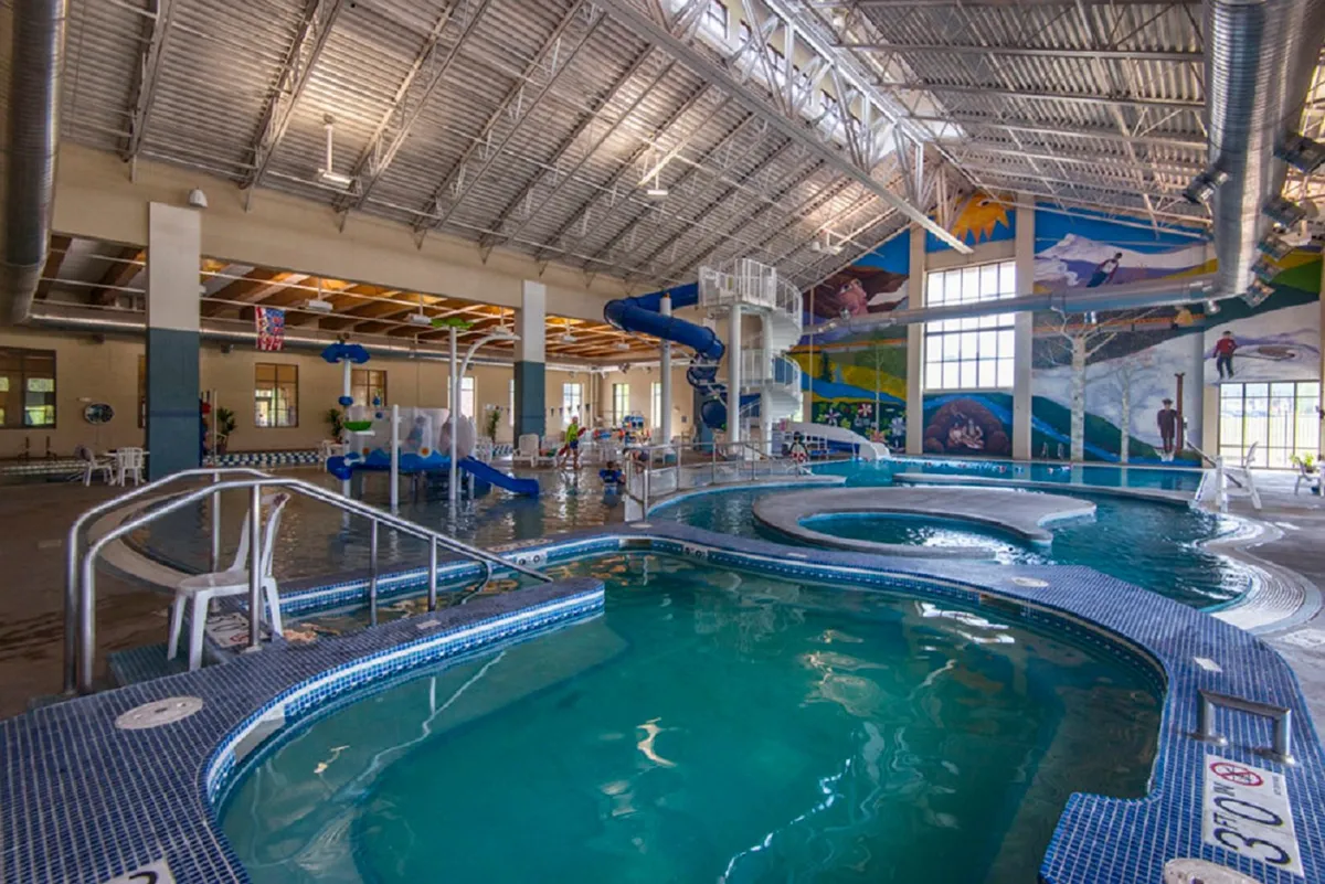 A bright indoor pool scene with a colorful waterslide, children playing, a mural on the wall, and a spa area. The joyful, energetic atmosphere highlights family-centric amenities available at the nearby Grand Park Rec Center.