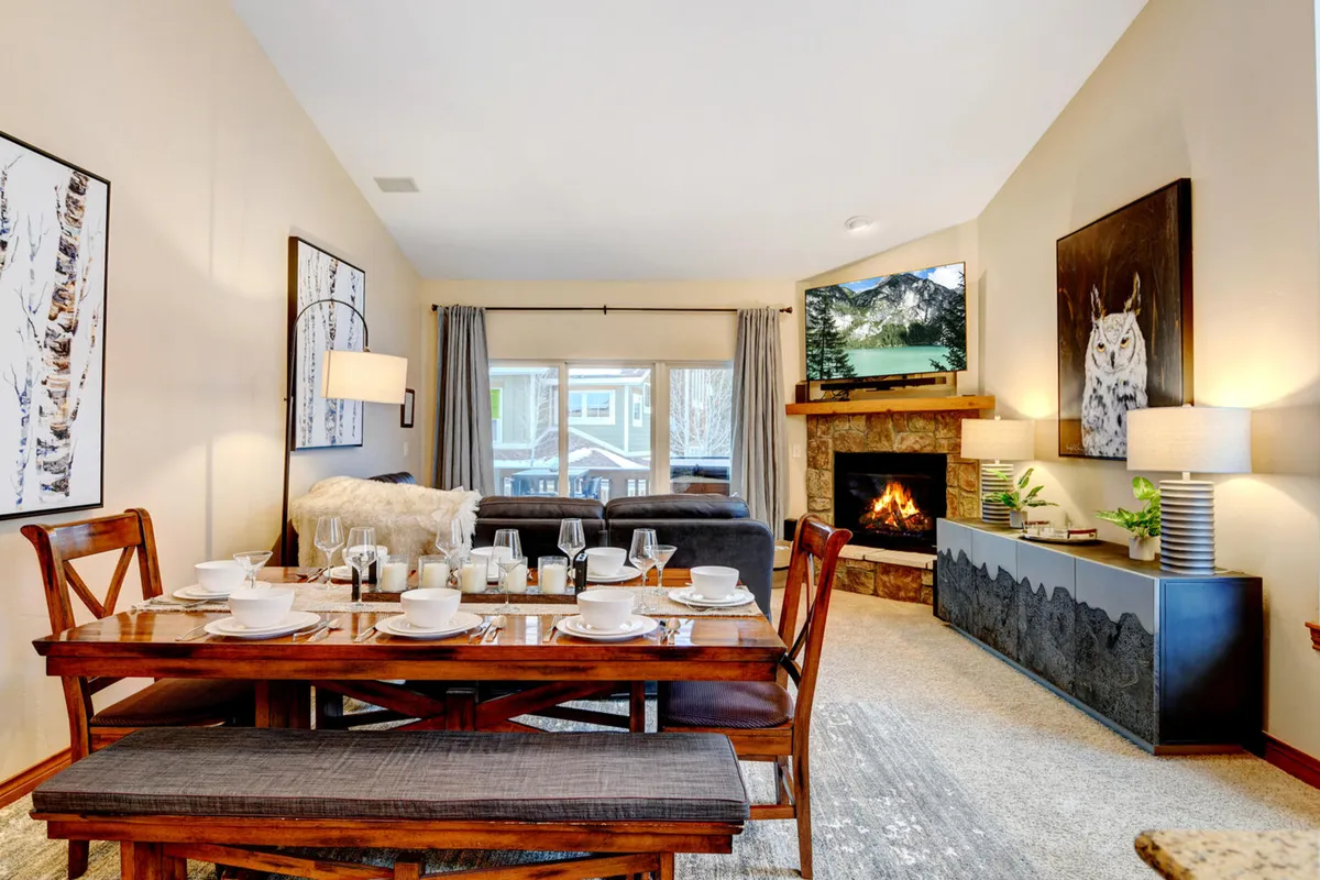 A warm and inviting living/dining area with a sofa, set dining table, fireplace, and TV. Artwork enhances the walls, and the arrangement fosters both cozy relaxation and lively group activities such as board games or meals.