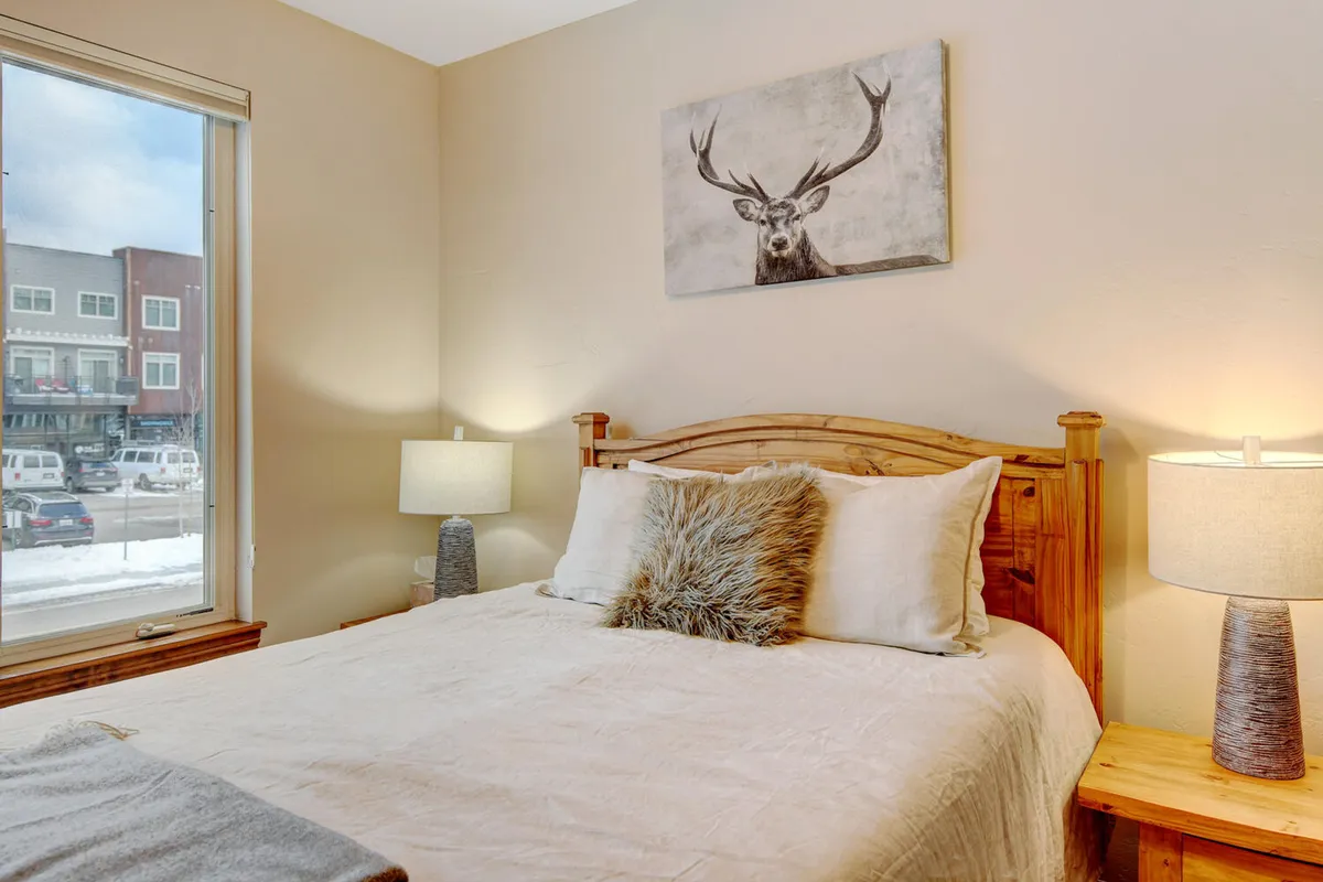 A serene master bedroom is showcased, featuring a plush king-sized bed adorned with premium bedding. Above the bed hangs striking deer artwork, while a large window lets in natural light and offers a view outside. Decorative pillows and a bedside lamp create a tranquil, restful atmosphere.