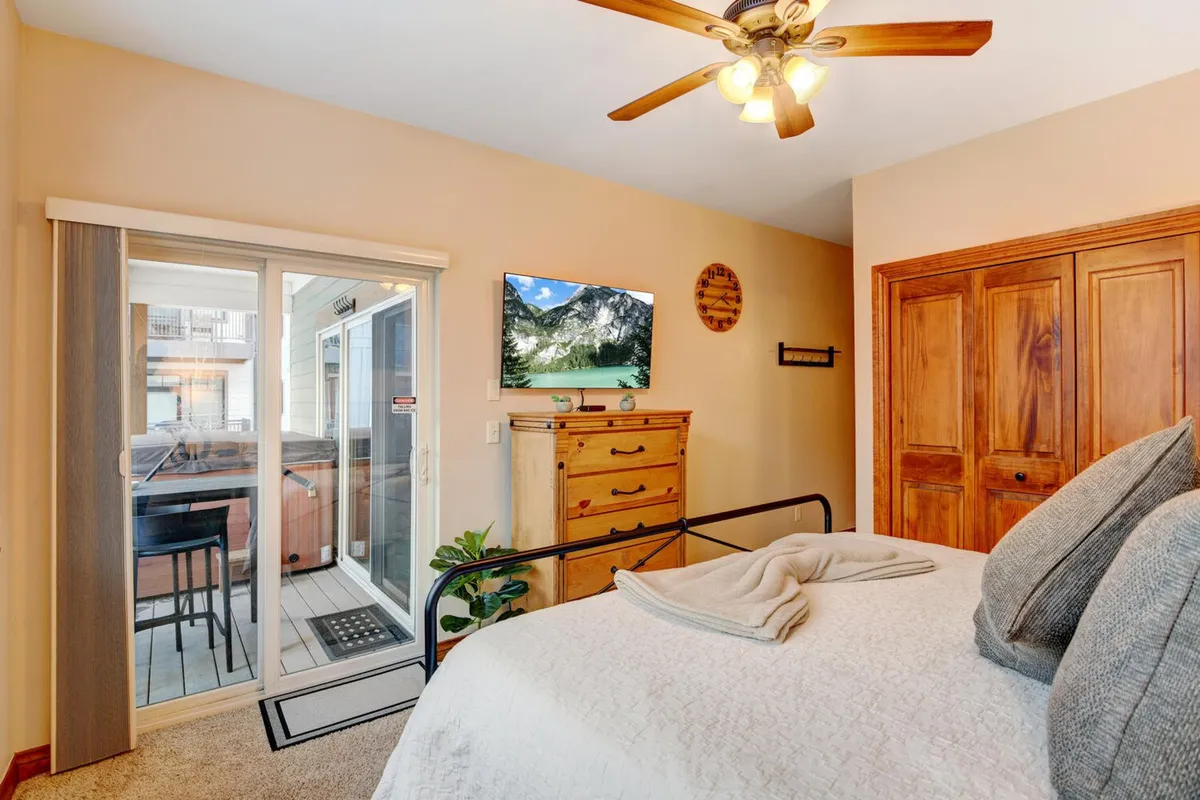 A serene bedroom with a large bed, sliding glass door opening to a balcony, a flat-screen television, and a dresser. The space is infused with natural light and offers direct access to a private outdoor space, implying relaxation after a day of adventure.