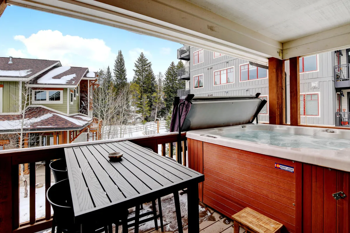 A tranquil exterior shot of a private hot tub on a wooden deck, overlooking tall trees and mountains. An outdoor table and wooden railing complete the inviting setup, suggesting relaxation and socializing after a day of activity.