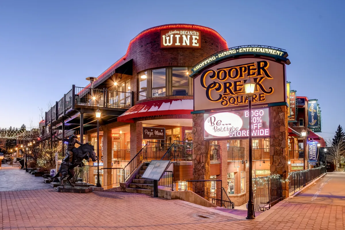 A vibrant exterior view of Cooper Creek Square, featuring its prominent sign, a wine shop entrance, outdoor seating, and a decorative horse statue. The image conveys a lively downtown atmosphere, ideal for shopping, dining, and seasonal festivals, showcasing the walkable, entertaining surroundings.