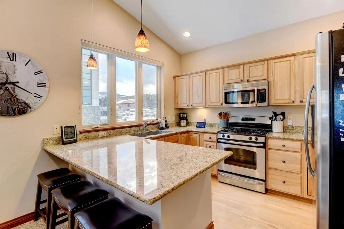 This image showcases a modern kitchen featuring granite countertops, stainless steel appliances, elegant wooden cabinets, warm pendant lighting, and large windows bringing in natural light. The scene captures a well-organized space perfect for preparing meals and entertaining guests.