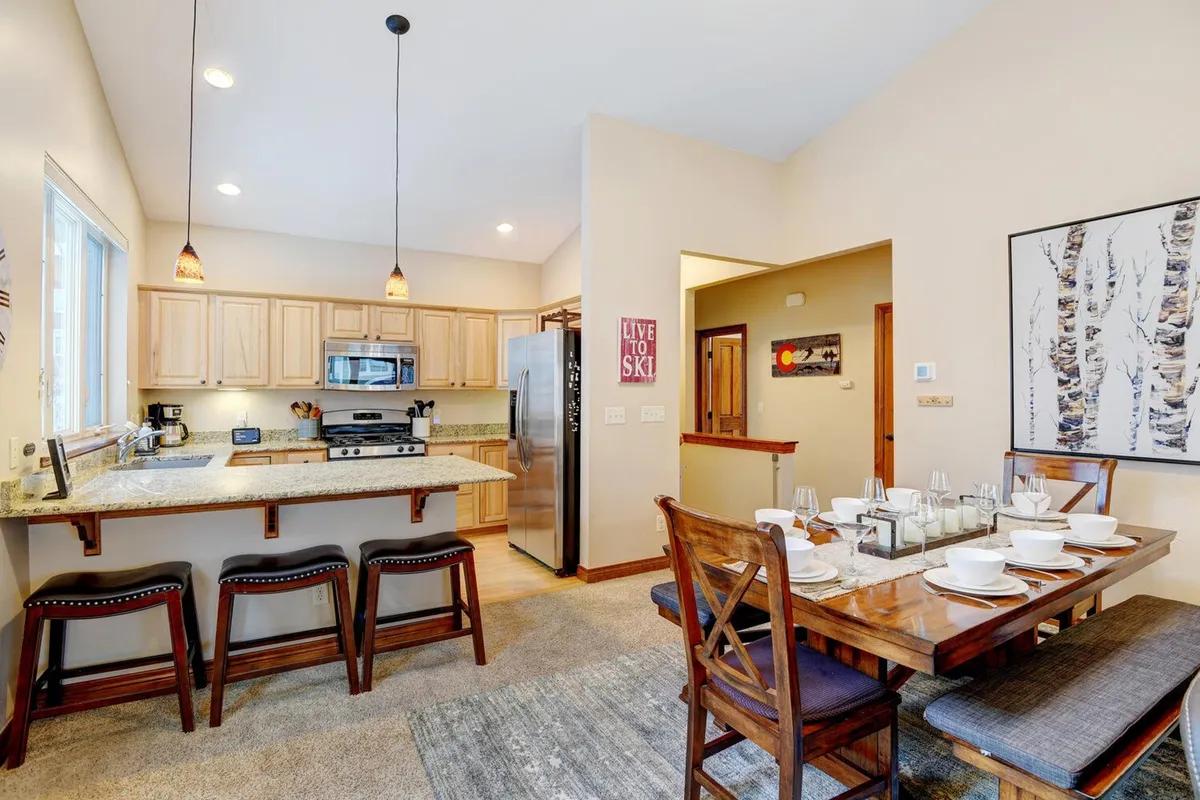 A dining area set for a meal, with a large wooden dining table, comfortable chairs, a kitchen island with bar stools, and visible kitchen appliances in the background. The space is warm, bright, and exudes a welcoming atmosphere for family meals or gatherings.