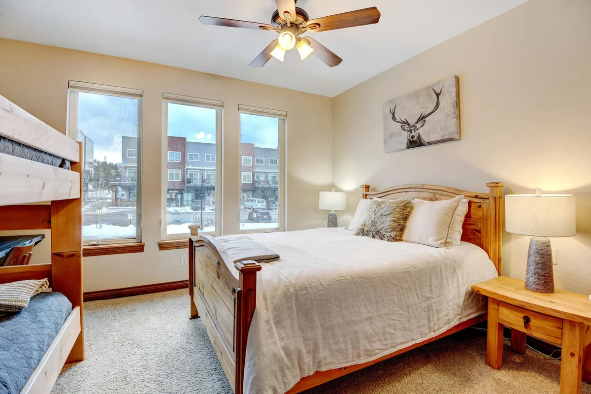 A cozy guest bedroom with a queen bed and a twin bunk bed, ideal for families or groups. Large windows brighten the room, and deer artwork above the bed adds a playful, welcoming touch. A nightstand with a lamp provides practical comfort.