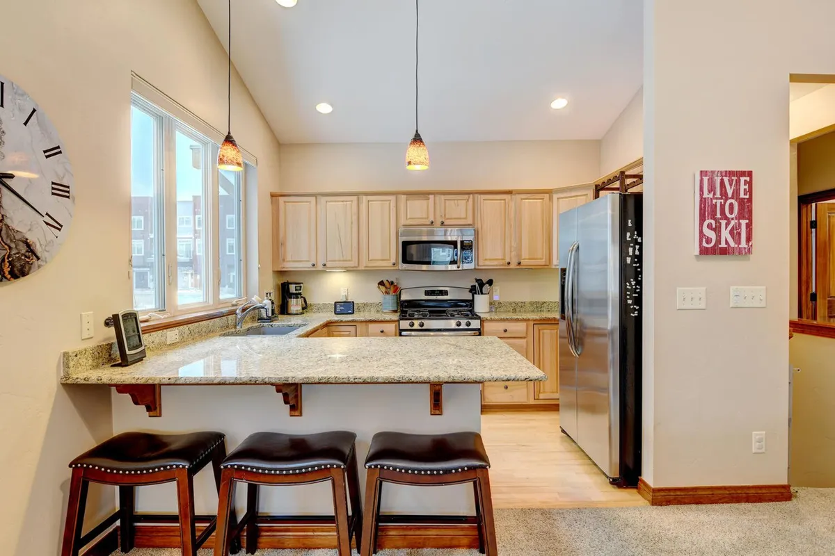 A cozy kitchen scene featuring a granite countertop with bar stools, modern kitchen appliances, and a visible wall clock. The layout is open, encouraging conversation and casual dining, and the atmosphere is bright and homey.