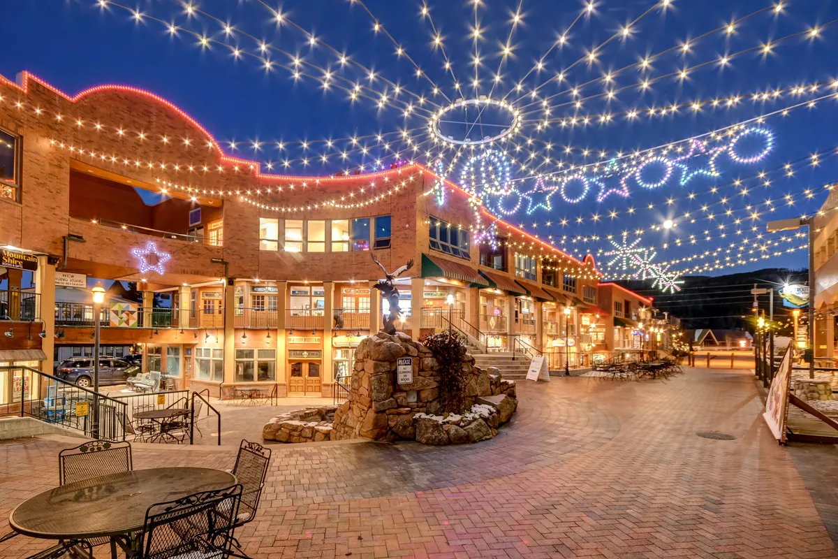 A lively exterior shot of downtown Winter Park at dusk, featuring overhead string lights, decorative stars, a charming stone fountain, and café tables ideal for socializing. The scene is bustling with people enjoying dining, shopping, and festive activities, capturing the spirit of the neighborhood.