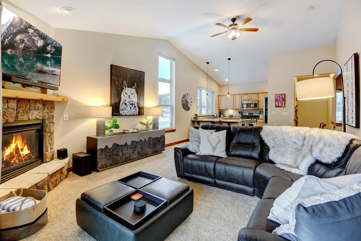 The image depicts a cozy and stylish living room with a modern fireplace as its centerpiece, flanked by a plush sofa, elegant coffee table, and tasteful artwork. The television is conveniently placed for entertainment, while large windows let in natural light and hint at snowy vistas outside, creating a warm and inviting atmosphere perfect for gatherings or relaxing after a day of mountain adventures.