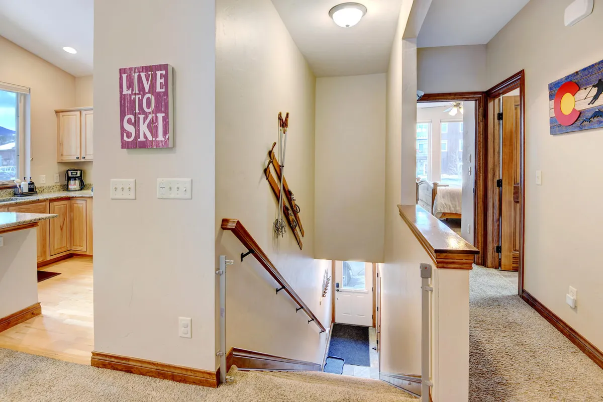 A cozy interior perspective showing a wooden staircase with ski-themed décor, a glimpse of the kitchen, and warm lighting. The space feels inviting and speaks to gathering after a day on the slopes, blending homey comfort with mountain charm.