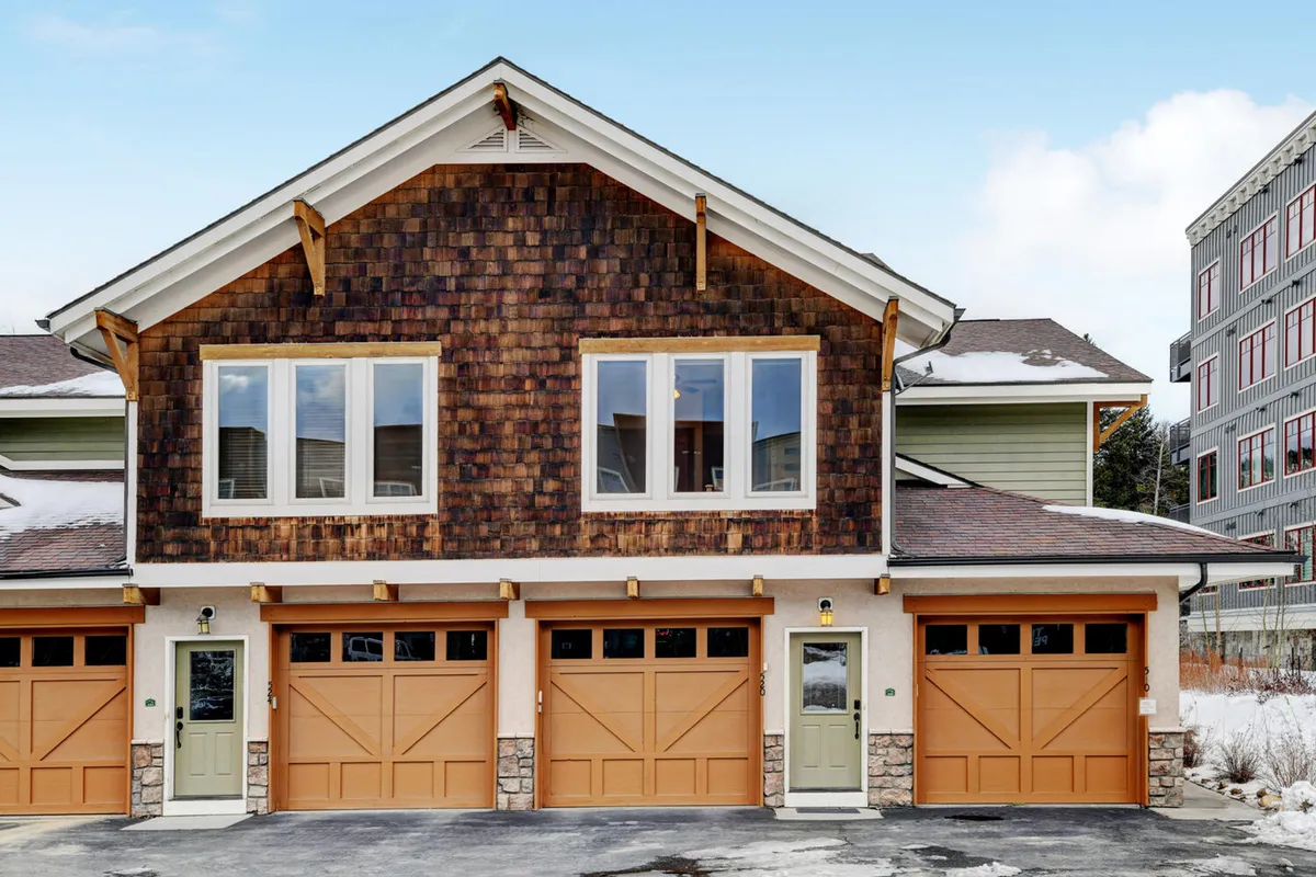 The image shows the exterior of the condo, featuring inviting wooden shingles, large windows, and garage doors. The entryway and porch area exude a welcoming vibe, hinting at both relaxation and practicality, with easy access for arriving with pets and outdoor gear.